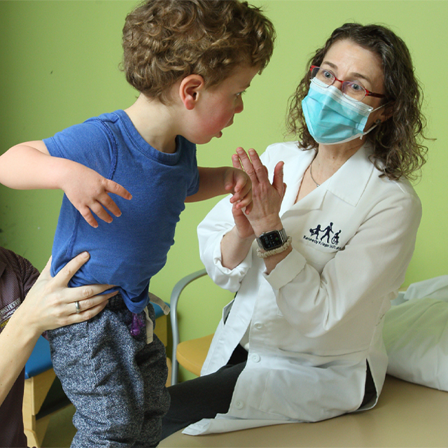 A toddler with curly hair in a blue shirt lifts his arms as he is held by a physical therapist, as Stacy Suskauer look on. 